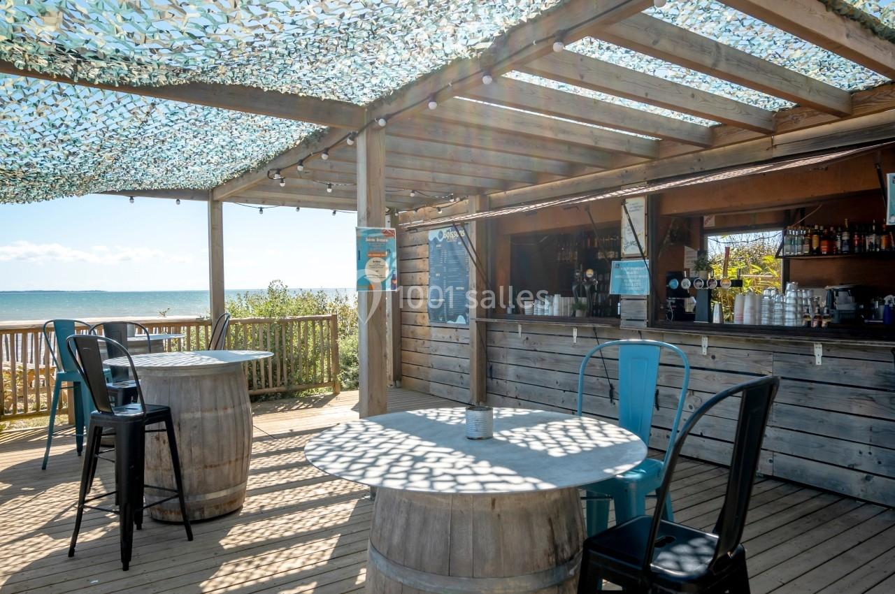 Terrasse en bois avec tables et chaises, bar en arrière-plan, vue sur la mer sous un toit en filet d'ombrage.