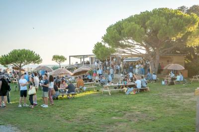 Groupe de personnes rassemblées autour de tables de pique-nique dans un espace extérieur avec des arbres et des parasols.