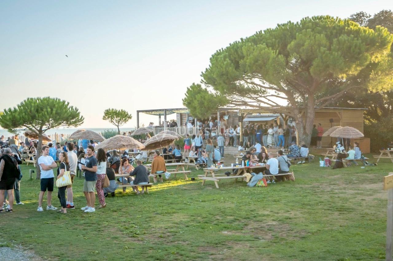 Groupe de personnes rassemblées autour de tables de pique-nique dans un espace extérieur avec des arbres et des parasols.