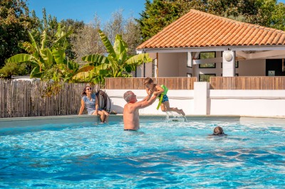 Groupe de personnes rassemblées autour de tables de pique-nique dans un espace extérieur avec des arbres et des parasols.