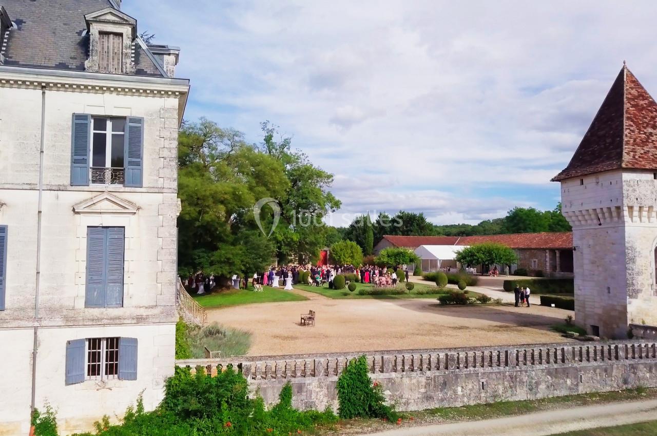 Vue d'une cour de château avec des invités rassemblés près d'un jardin, entourée de bâtiments anciens et d'arbres.