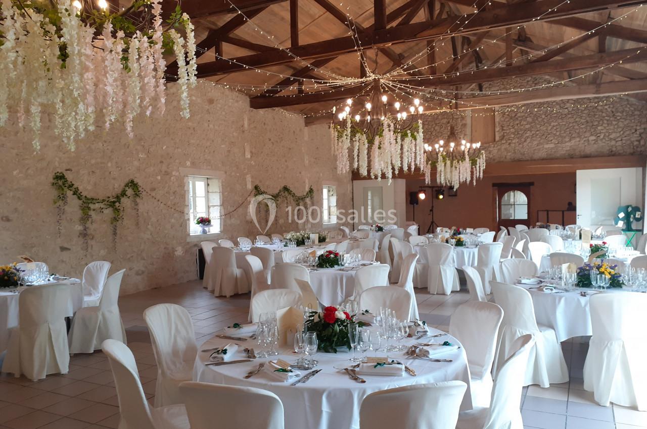 Salle de réception décorée avec des tables rondes dressées, des chaises blanches et des guirlandes lumineuses suspendues.
