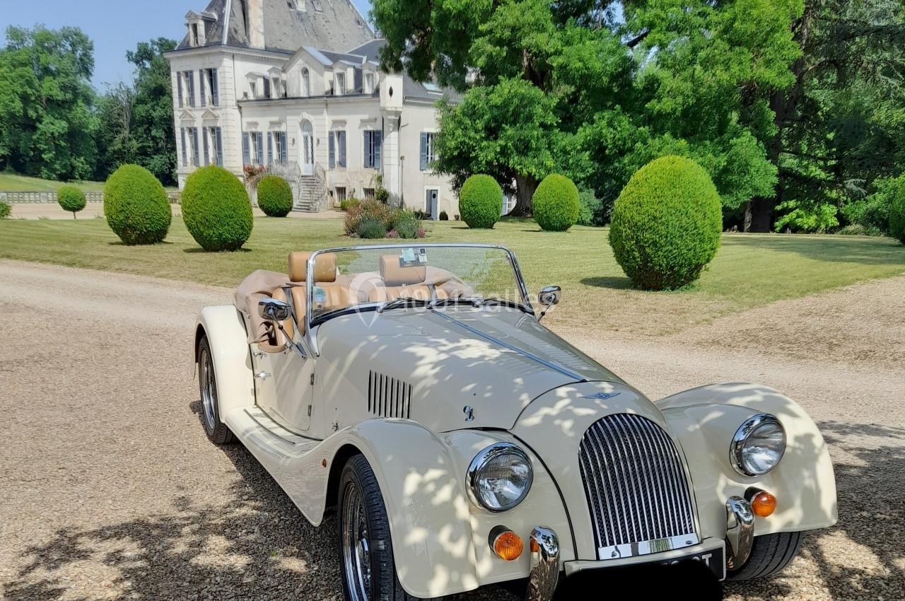 Voiture de collection décapotable beige stationnée sur une allée gravillonnée devant un manoir entouré de verdure.