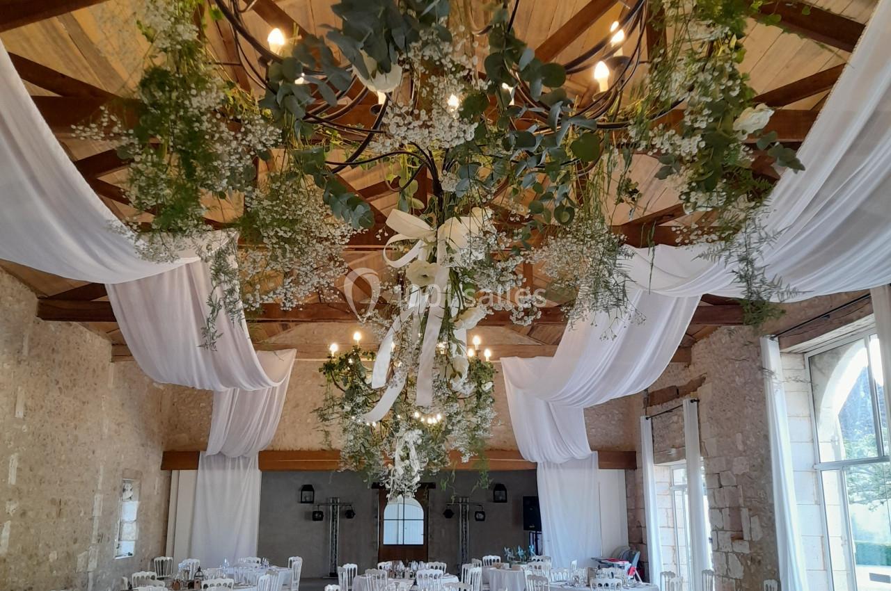 Salle de réception décorée avec des tables dressées, des chaises blanches et un plafond orné de guirlandes végétales.