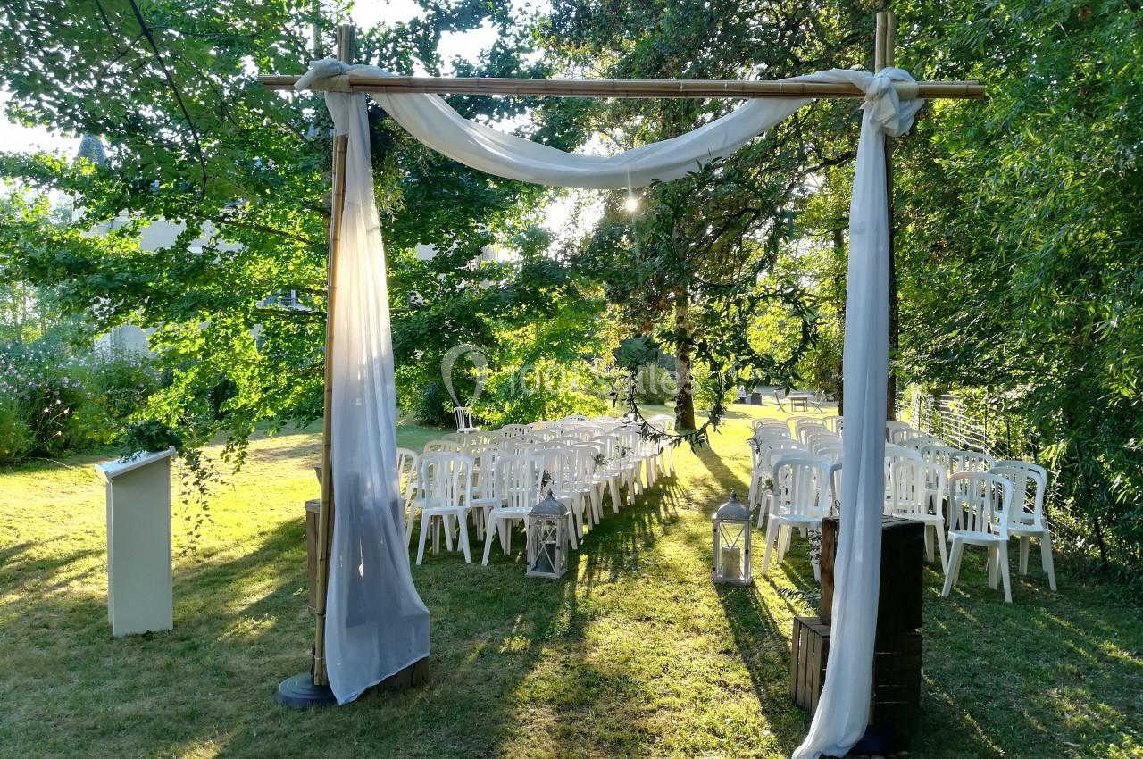 Arche décorée de voilages blancs devant des rangées de chaises blanches disposées en extérieur, sous des arbres.