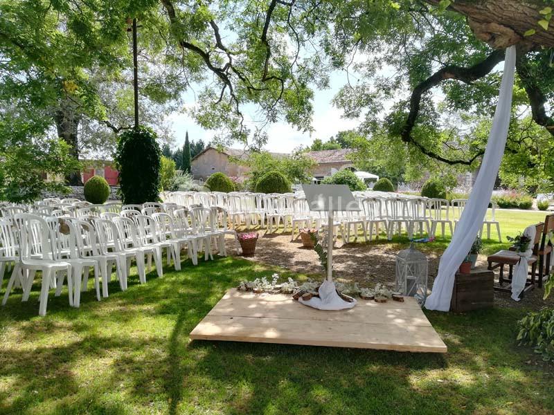 Chaises blanches disposées en extérieur sous des arbres, face à une petite estrade décorée pour une cérémonie.