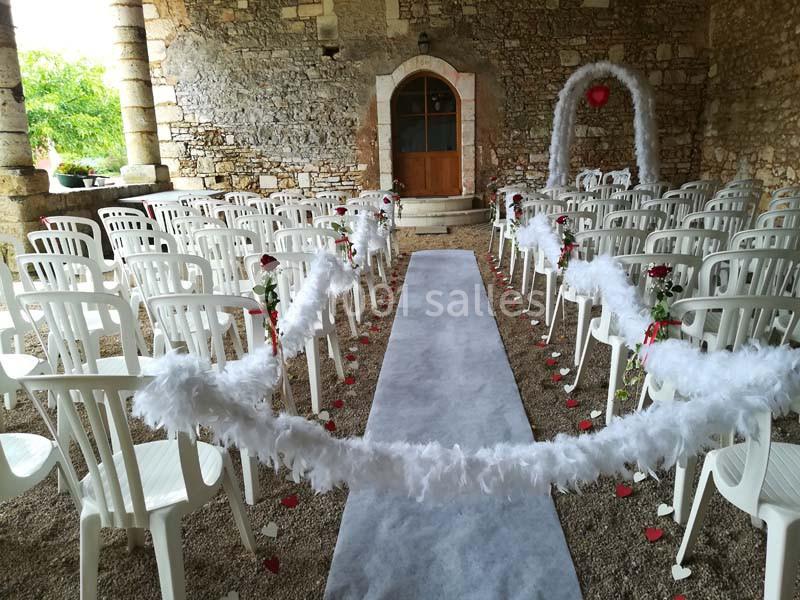 Allée centrale décorée de plumes blanches et de roses rouges, entourée de chaises blanches dans une salle en pierre.
