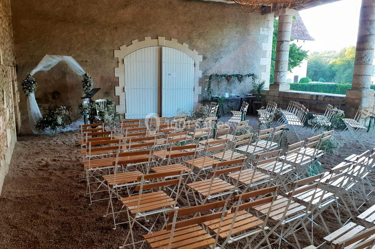 Chaises en bois disposées pour une cérémonie dans une cour couverte avec arche florale et vue sur un jardin.