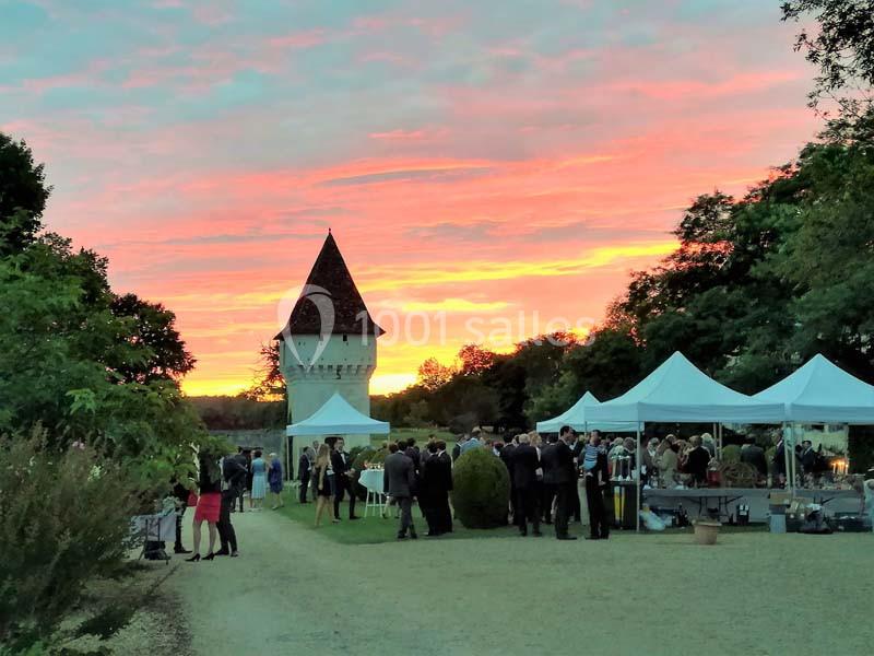 Groupe de personnes rassemblées près de tentes blanches dans un jardin au coucher du soleil, avec une tour en arrière-plan.