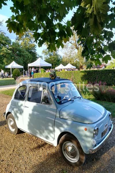 Petite voiture vintage bleu clair garée sur un chemin gravillonné, entourée de verdure et de tentes blanches en arrière-plan.