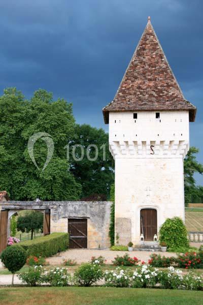 Tour en pierre avec toit en tuiles, entourée de jardins fleuris et d'arbres sous un ciel nuageux.