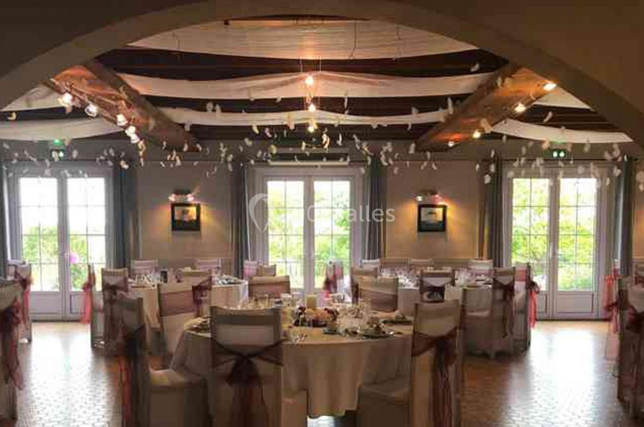 Salle de réception décorée avec des tables rondes dressées, chaises ornées de rubans et lumière naturelle.