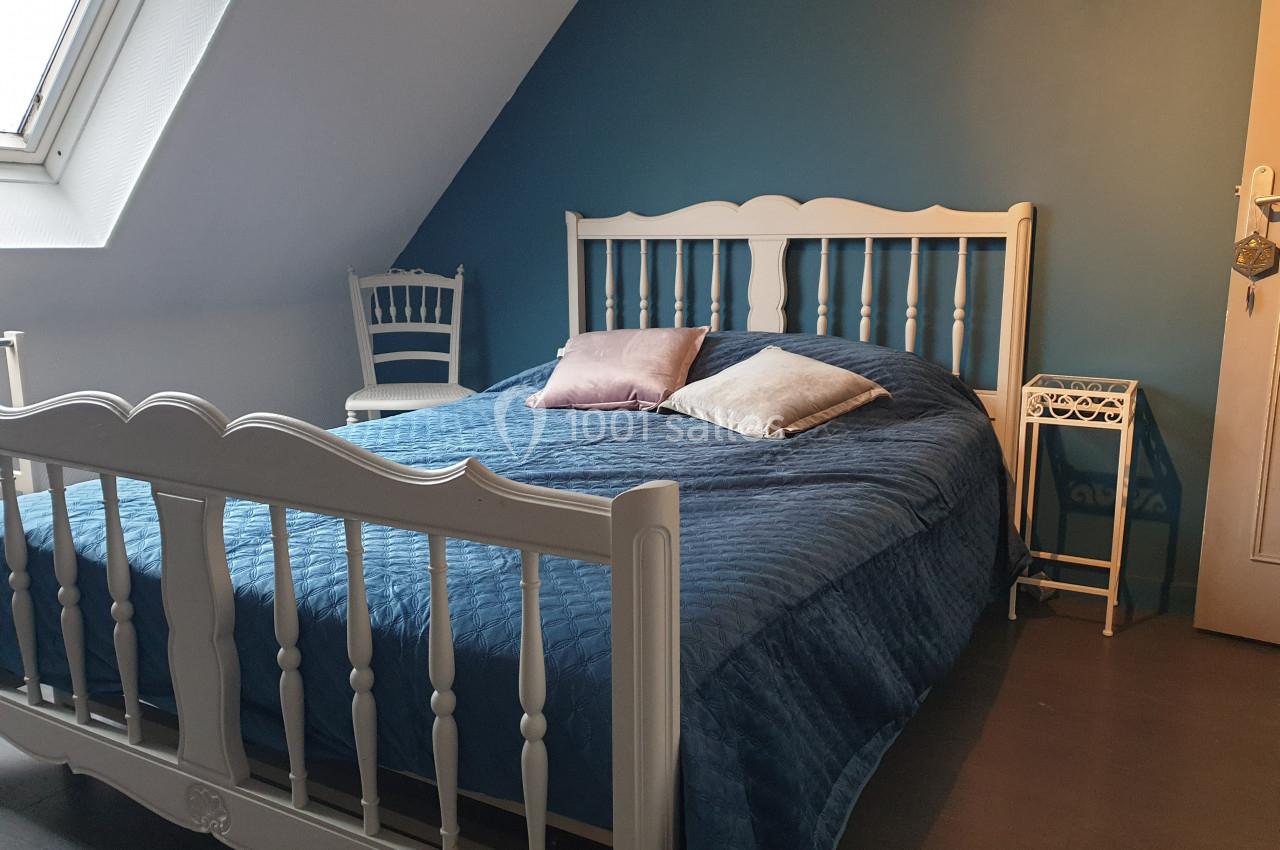 Lit double avec couvre-lit bleu, deux coussins, encadré par des meubles blancs dans une chambre mansardée.