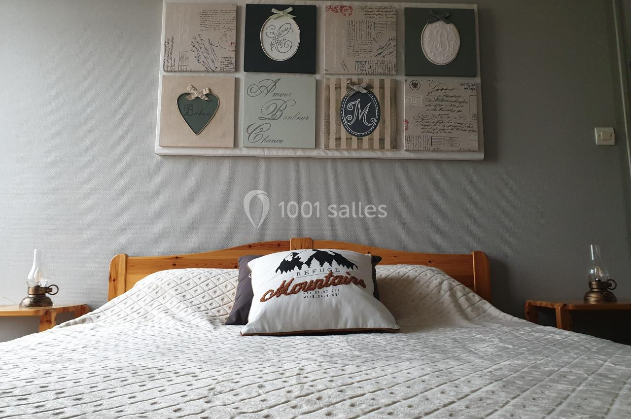 Lit double avec couvre-lit beige, coussin décoratif ’Mountains’ et tableau mural avec motifs variés en arrière-plan.