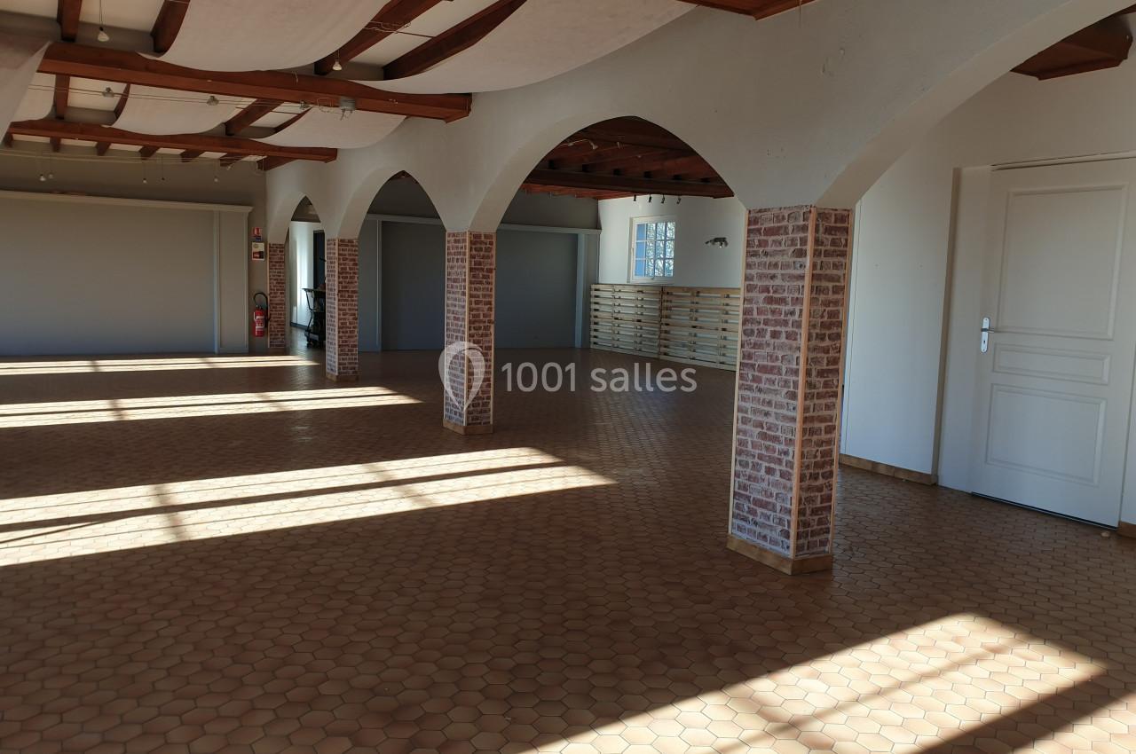 Salle spacieuse avec sol en carreaux de terre cuite, murs clairs, poutres apparentes et piliers en briques.