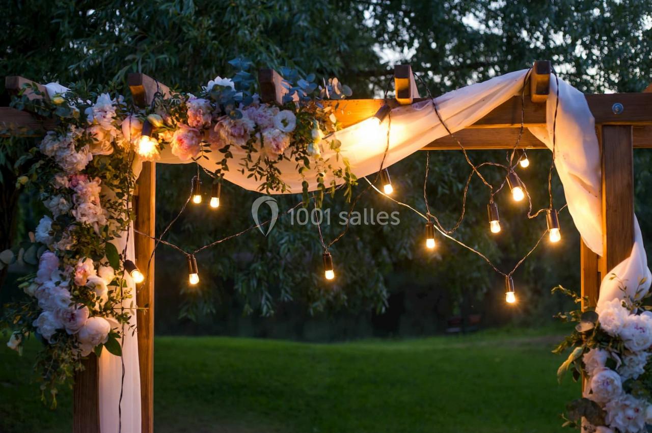 Pergola en bois décorée de guirlandes lumineuses, de fleurs et de tissu blanc, dans un jardin au crépuscule.