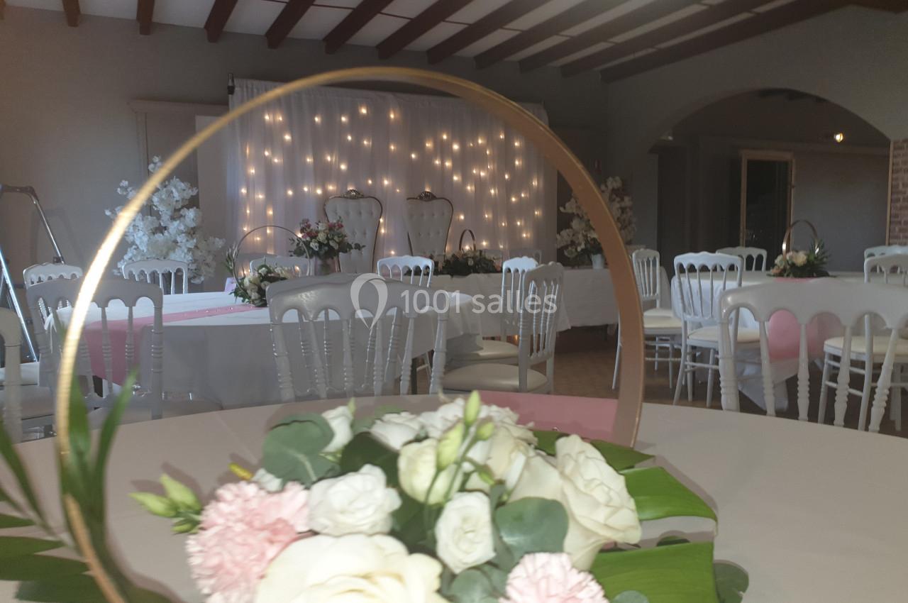 Salle de réception décorée avec des tables rondes, des chaises blanches et des fleurs, éclairée par des guirlandes…