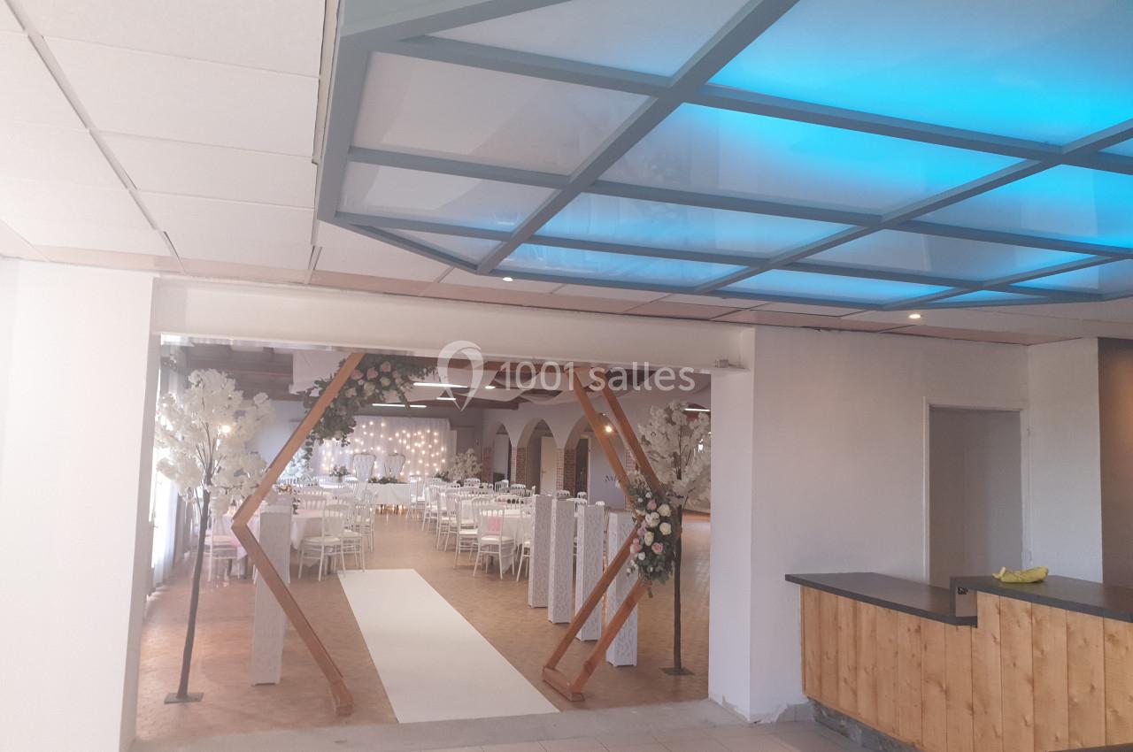 Salle de réception décorée avec des tables dressées, des chaises blanches et une arche fleurie à l'entrée.