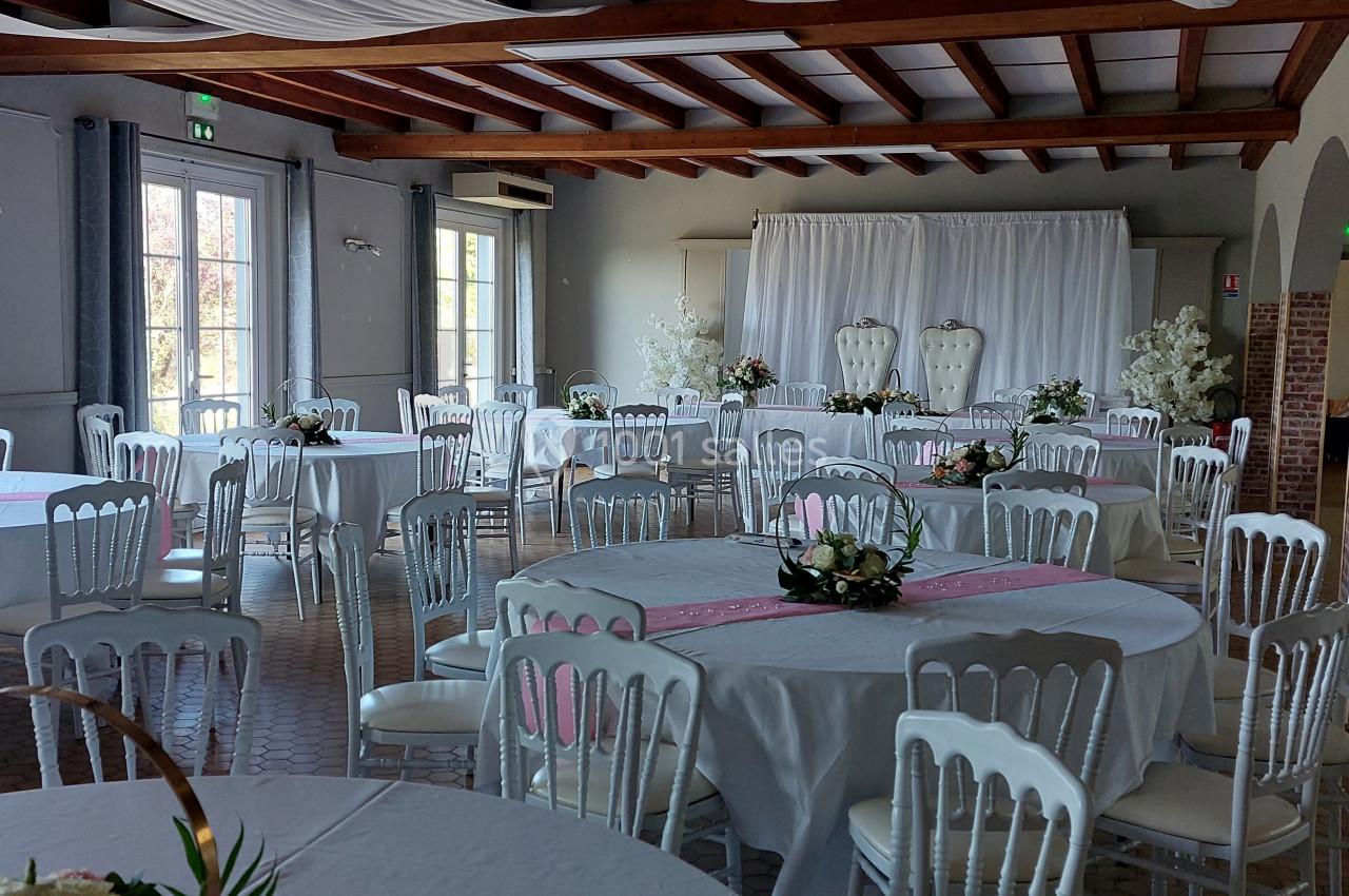 Salle de réception lumineuse avec tables rondes dressées, décorations florales et deux fauteuils blancs en arrière-plan.