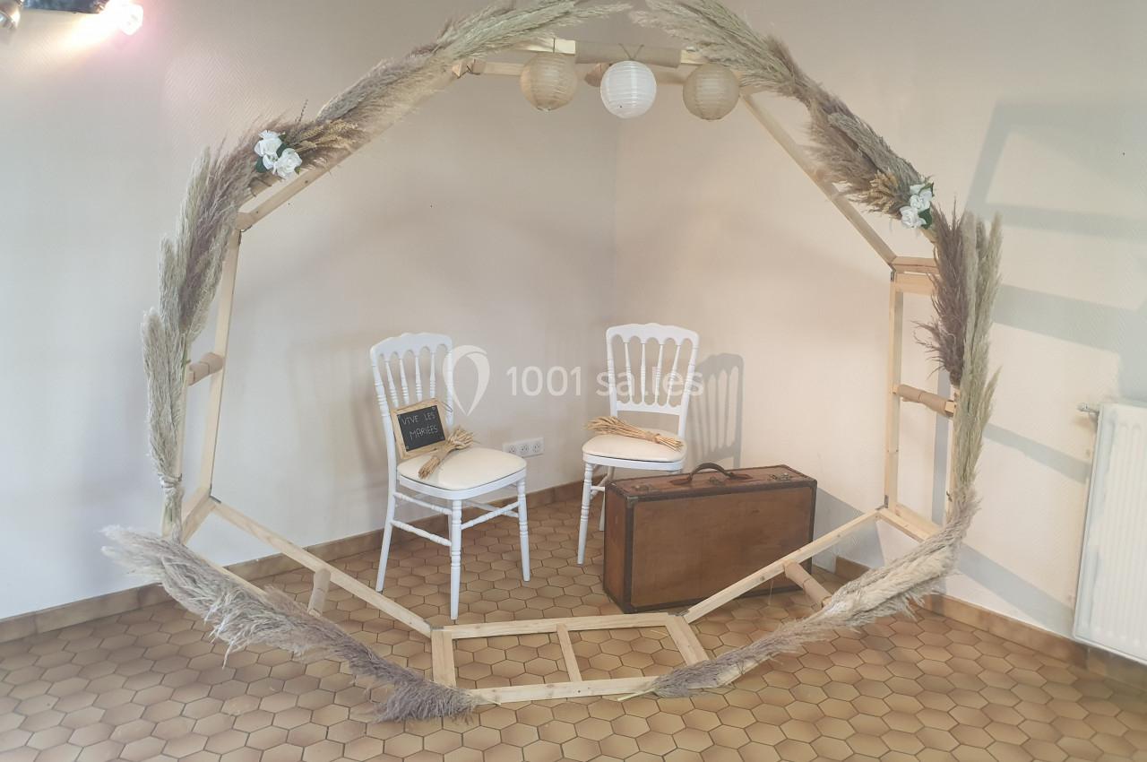 Structure circulaire en bois décorée de pampas et lanternes, avec deux chaises blanches et une malle en arrière-plan.