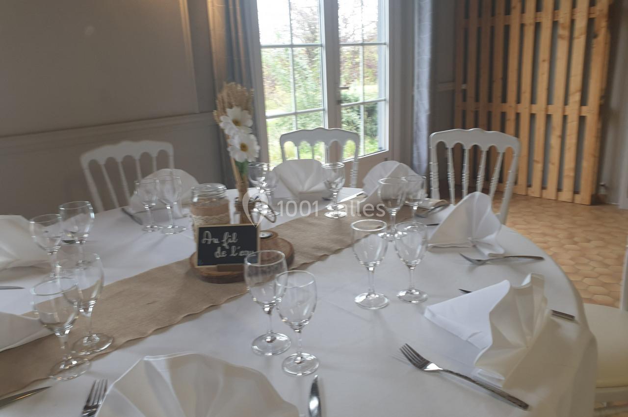Table ronde dressée avec nappes blanches, serviettes pliées, verres et couverts, décorée de fleurs et d'une ardoise.