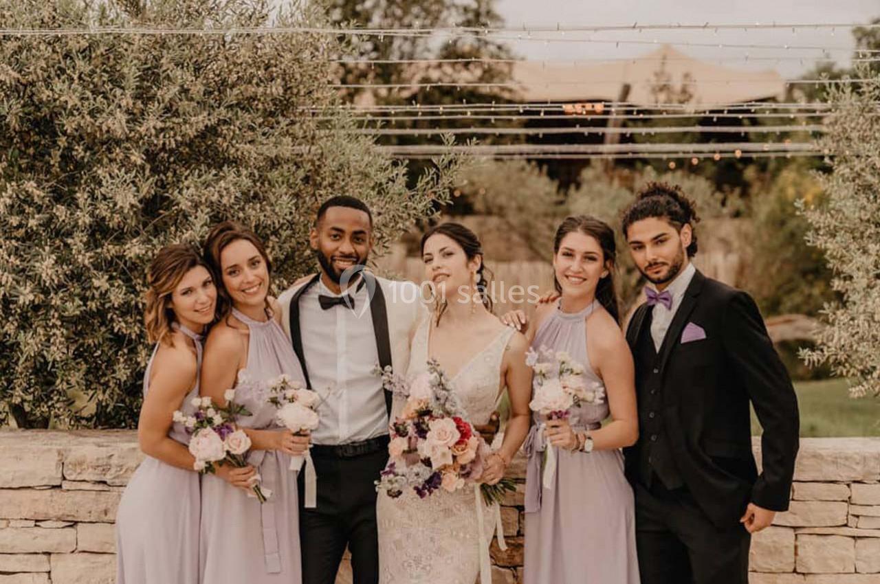 Un couple de mariés pose avec quatre invités en tenue élégante, devant un décor extérieur avec des guirlandes lumineuses.