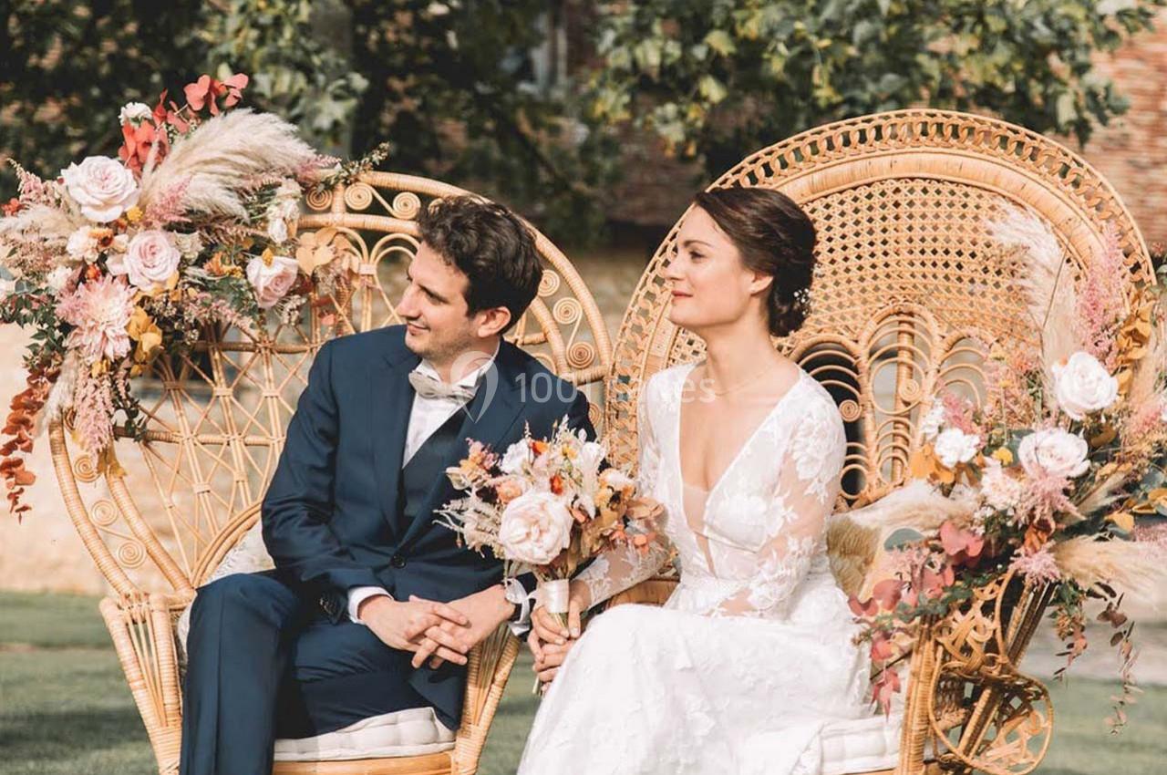 Un couple en tenue de mariage est assis sur des fauteuils en rotin décorés de fleurs, en extérieur.