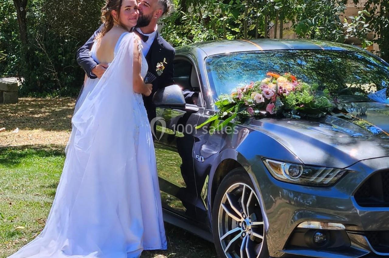Un couple en tenue de mariage pose près d'une voiture décorée de fleurs dans un jardin verdoyant.