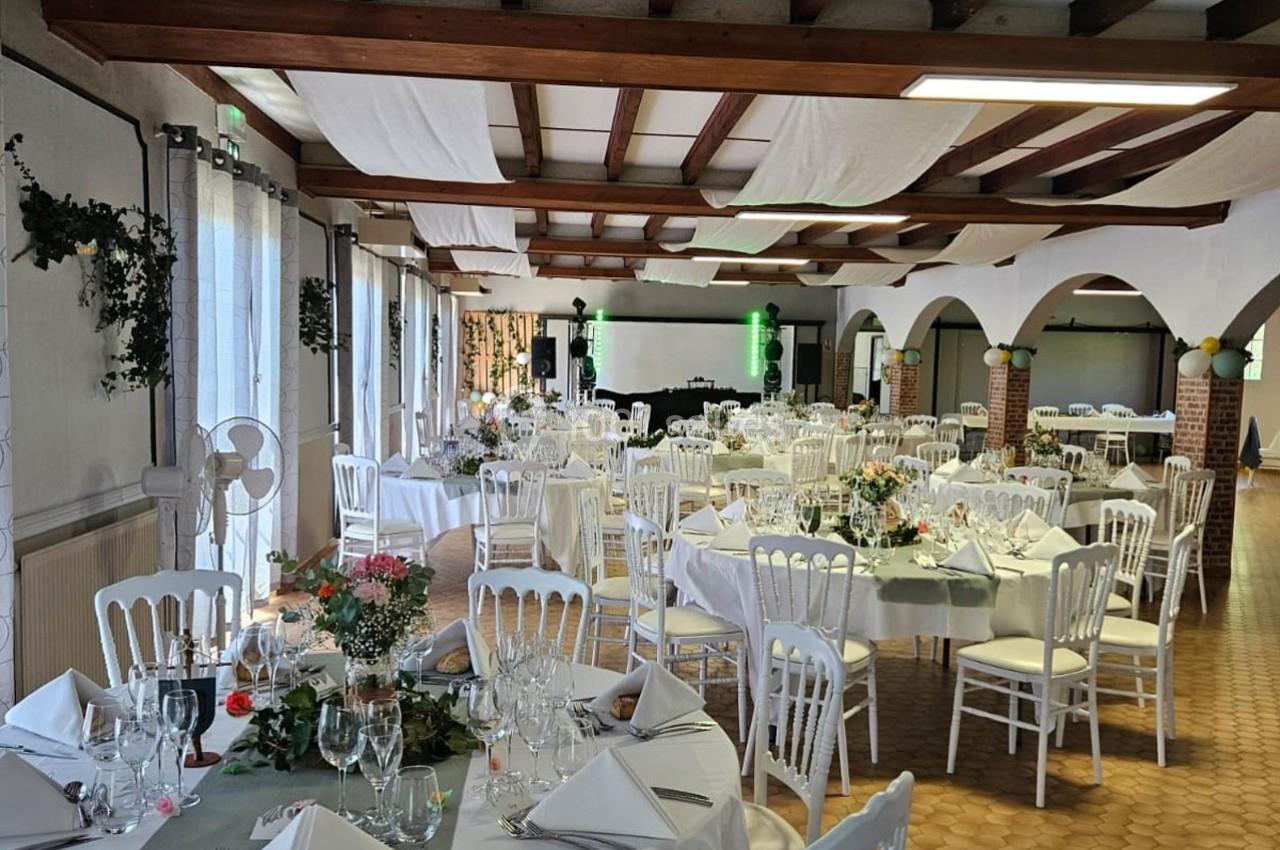 Salle de réception décorée avec des tables dressées, chaises blanches et ornements floraux, prête pour un événement.