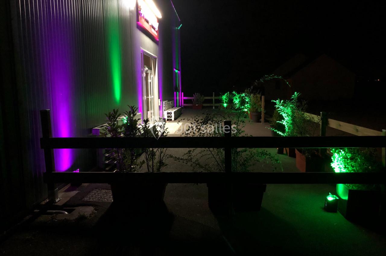 Extérieur d'un bâtiment éclairé par des lumières violettes et vertes, avec des plantes en pot et une clôture en bois.