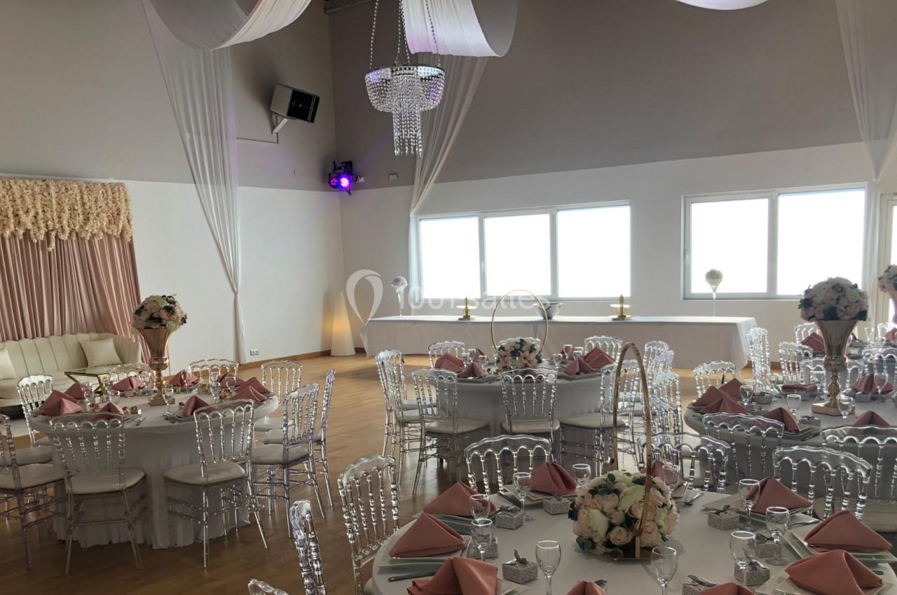 Salle de réception décorée avec des tables rondes, nappes blanches, serviettes roses et chaises transparentes.