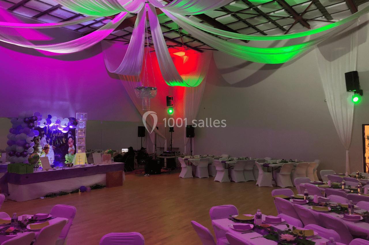 Salle de réception décorée avec des drapés blancs au plafond, éclairages colorés et tables dressées pour un événement.