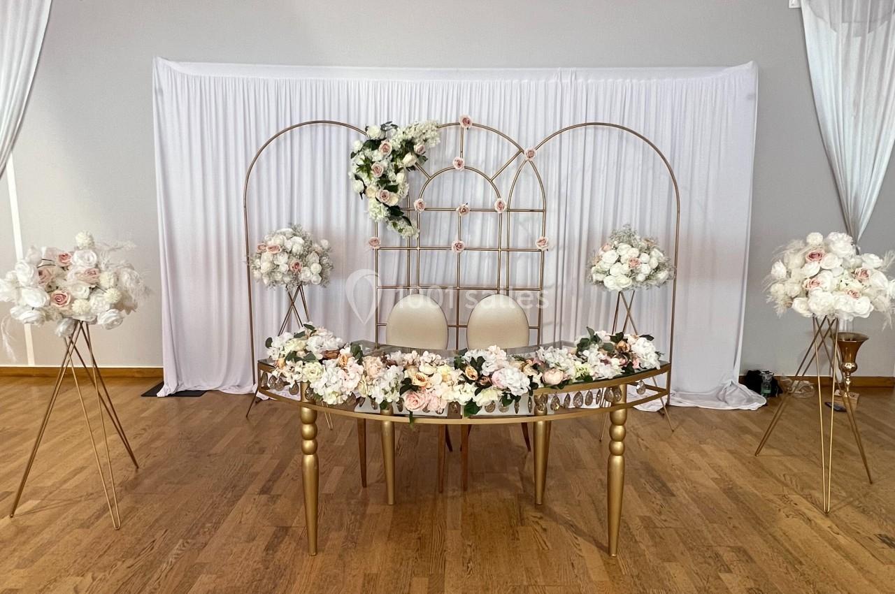 Décoration de mariage avec table ornée de fleurs blanches et roses, chaises blanches et arche métallique dorée.