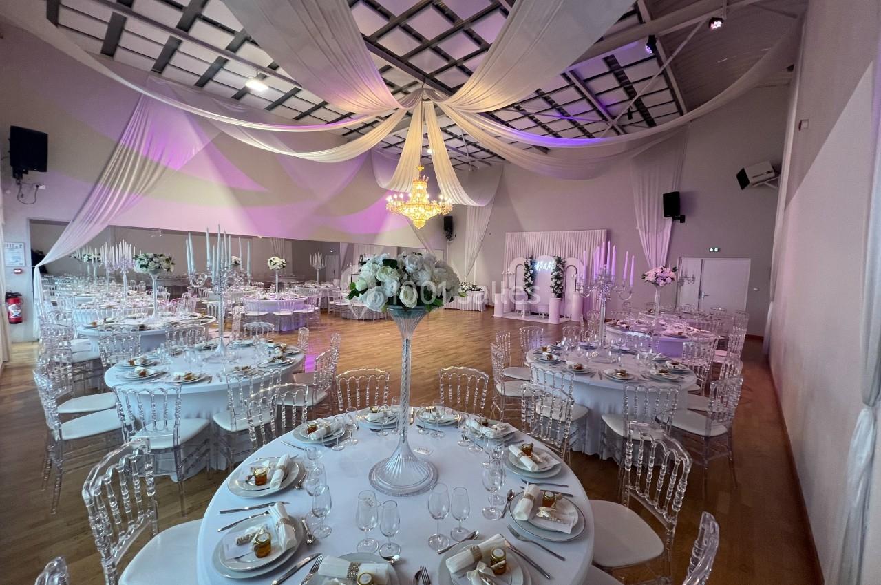 Salle de réception décorée avec des tables rondes dressées, des chaises transparentes et des drapés suspendus au plafond.