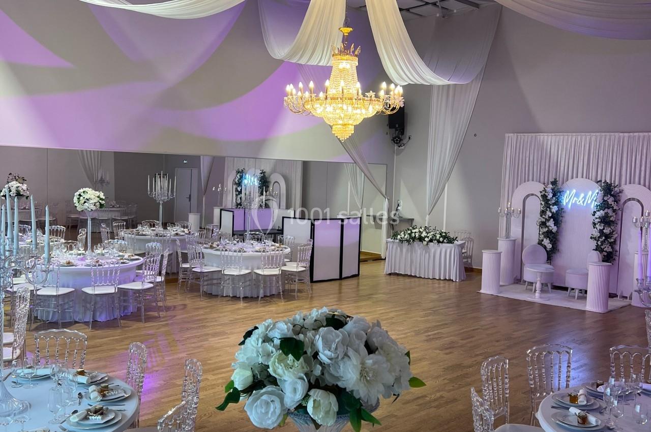Salle de réception élégante avec tables décorées, chaises transparentes, lustre central et drapés suspendus au plafond.
