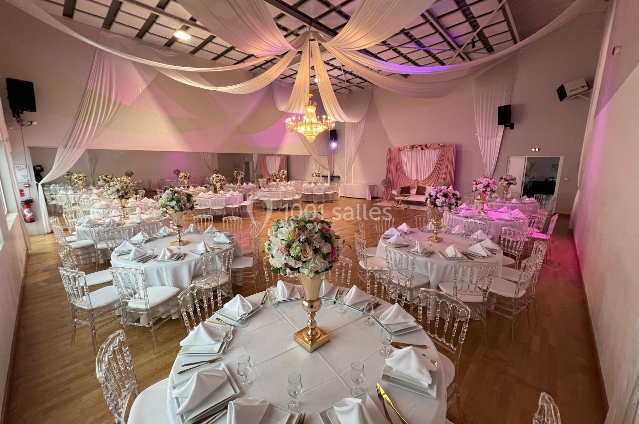 Salle de réception décorée pour un mariage, avec tables rondes dressées, chaises transparentes et plafonds ornés de drapés.