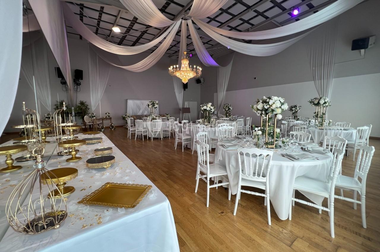 Salle de réception décorée avec des tables rondes, des chaises blanches, des fleurs et des drapés suspendus au plafond.