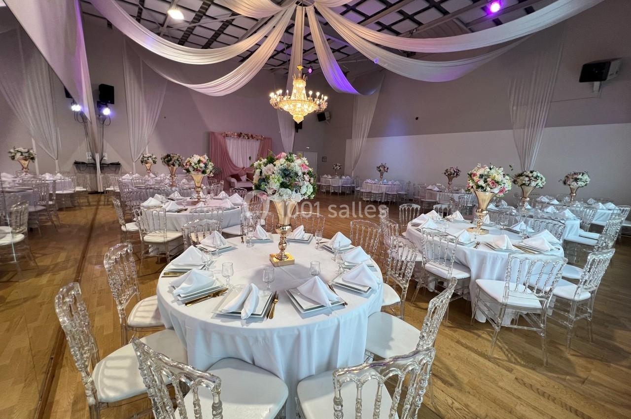 Salle de réception décorée avec des tables rondes, nappes blanches, chaises transparentes et un lustre central.