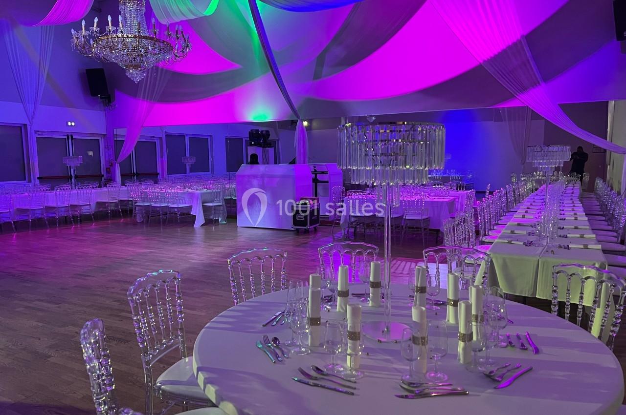 Salle de réception décorée avec des tables dressées, des chaises transparentes et un éclairage coloré.