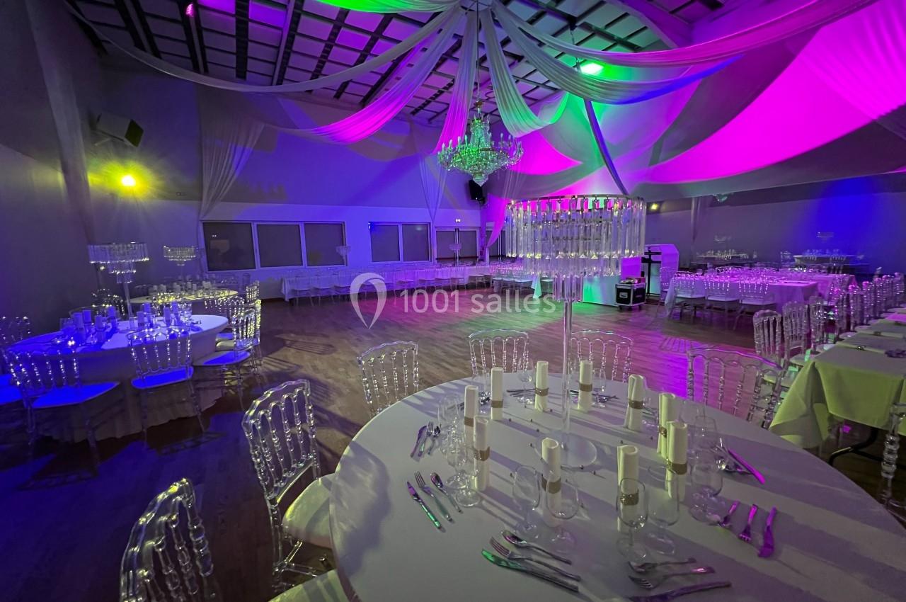 Salle de réception décorée avec des tables dressées, des chaises transparentes et un éclairage coloré.