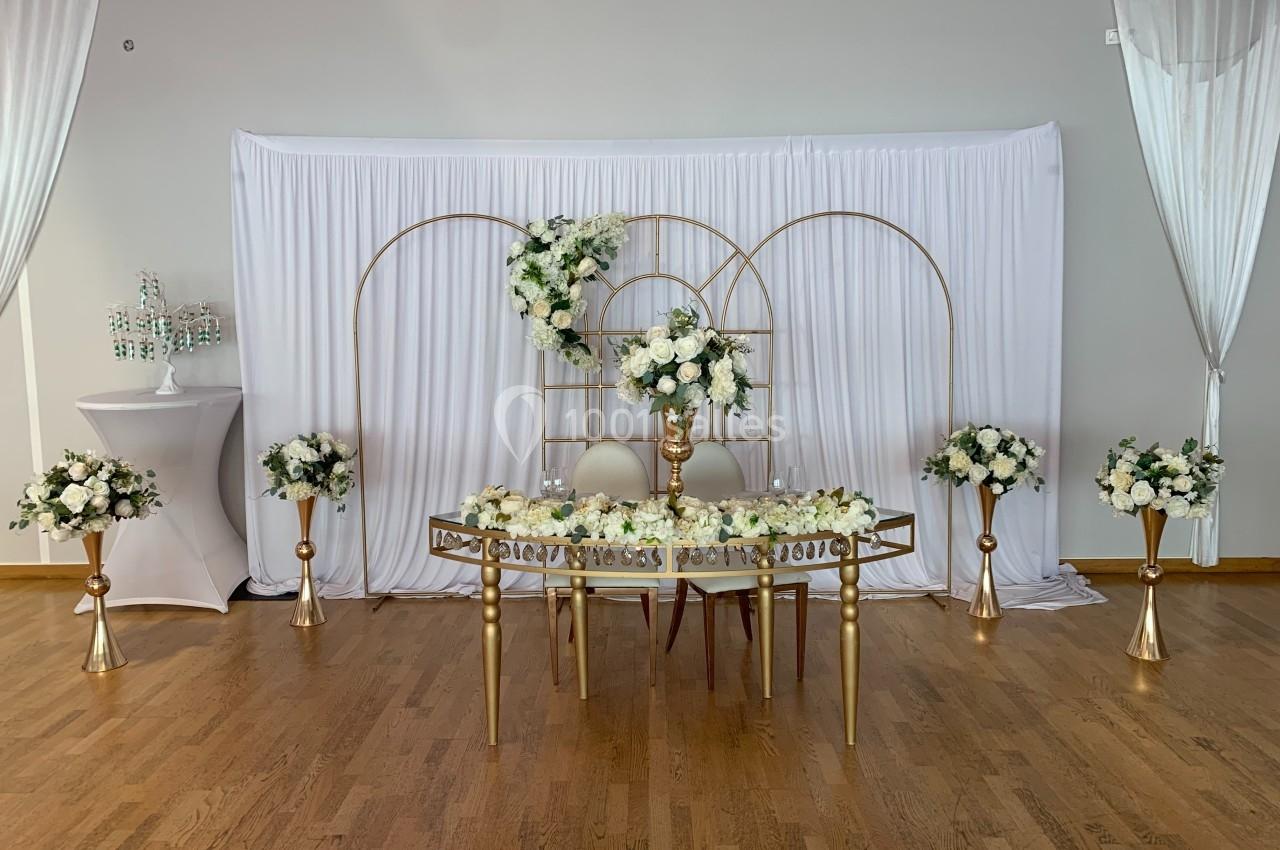 Décoration de mariage avec table dorée, fleurs blanches et rideaux blancs en arrière-plan.