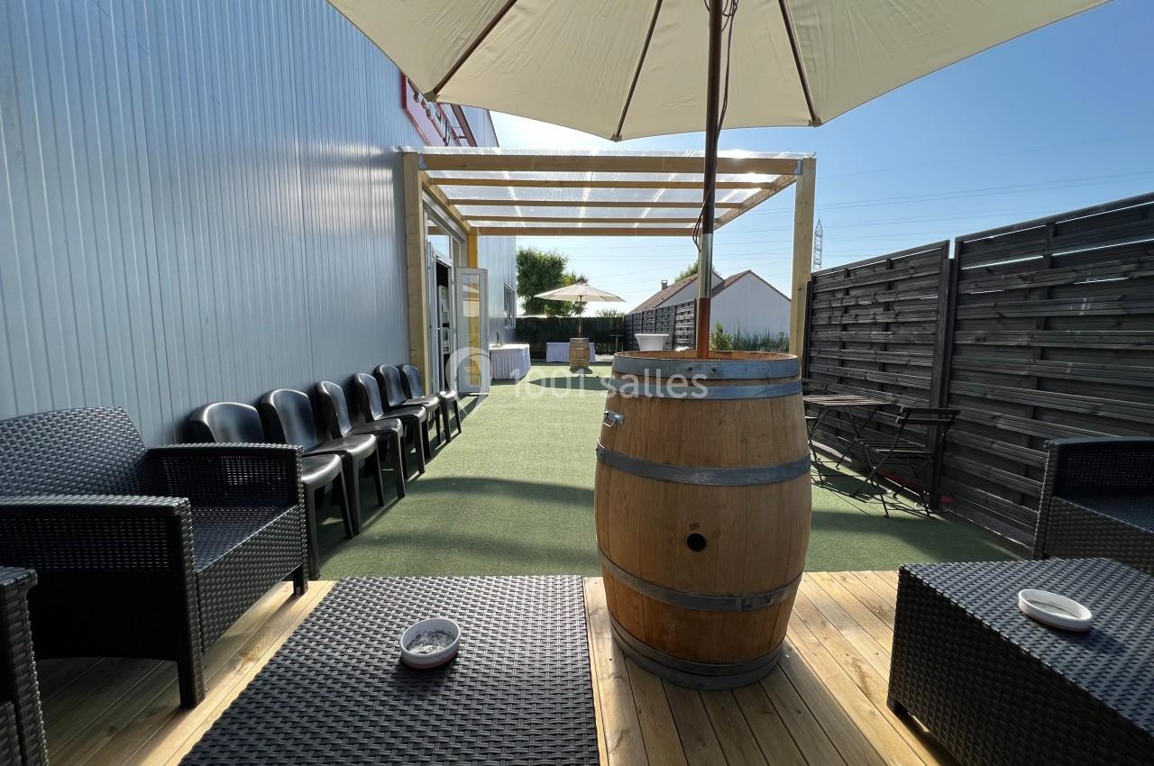 Terrasse extérieure avec mobilier en rotin, tonneau en bois, parasol et pergola, entourée de panneaux en bois.