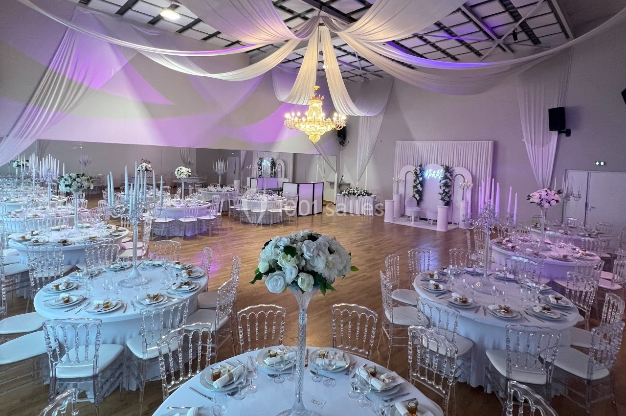 Salle de réception décorée avec des tables rondes, des chaises transparentes, un lustre central et des drapés au plafond.