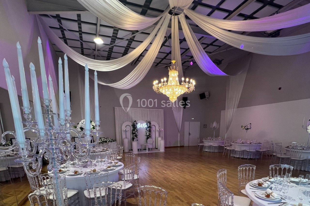 Salle de réception décorée avec des chandeliers, des tables rondes dressées et un grand lustre central.