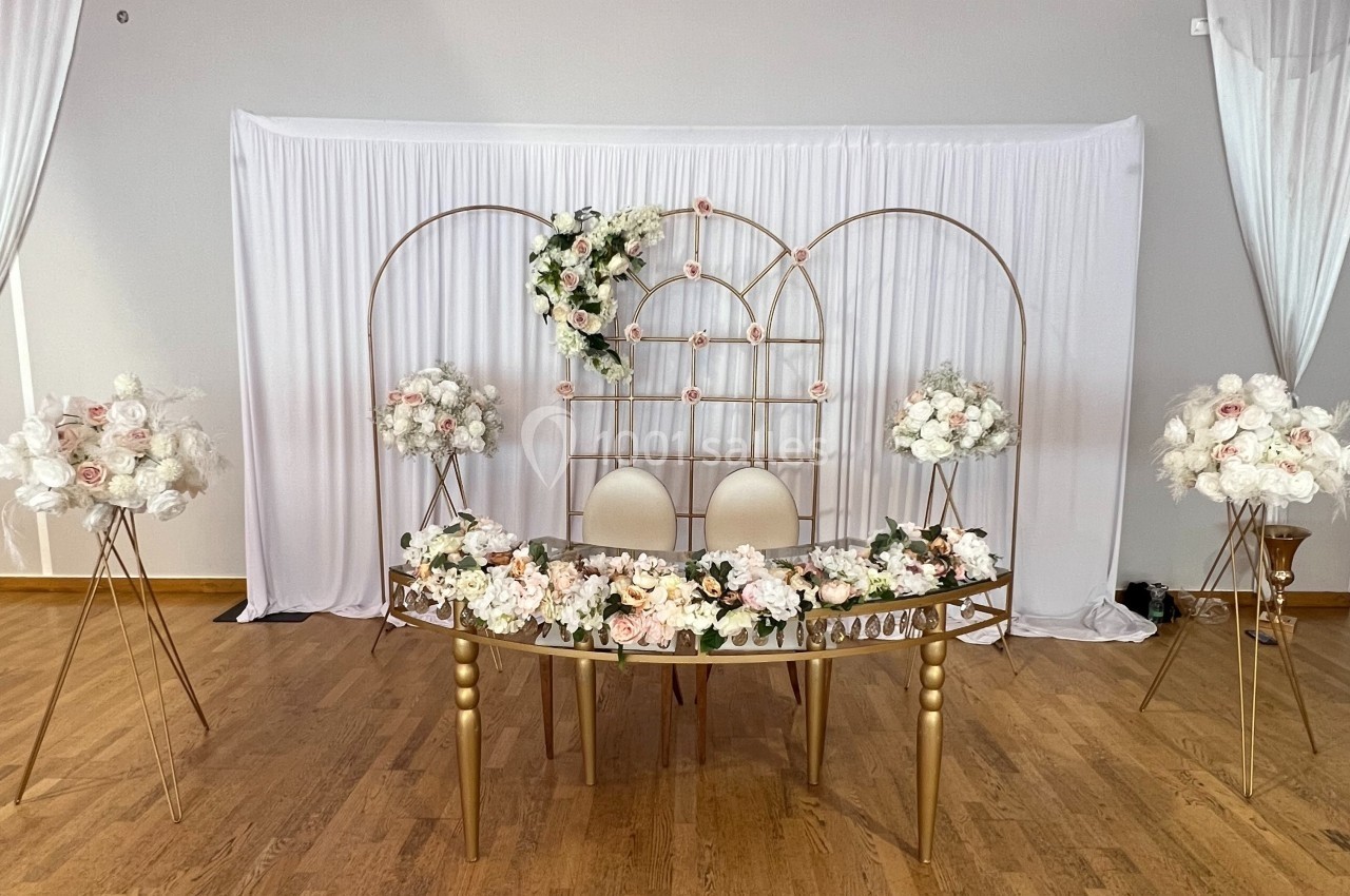 Décoration de table de mariage avec fleurs blanches et roses, chaises blanches et arche dorée devant un rideau blanc.