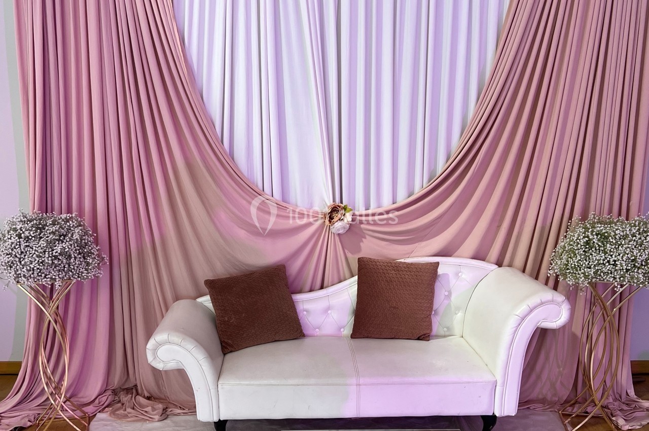 Canapé blanc avec coussins marron devant un rideau rose drapé, encadré par deux bouquets de gypsophiles.