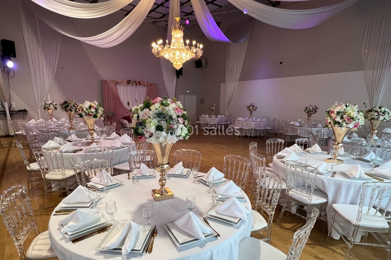 Salle de réception décorée avec des tables rondes dressées, des chaises transparentes et des arrangements floraux élégants.