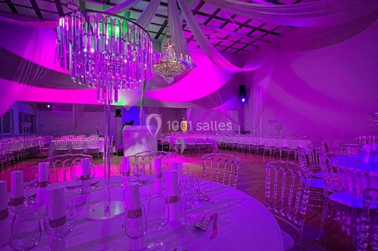Salle de réception décorée avec des tables dressées, des chaises transparentes et un éclairage coloré violet et vert.