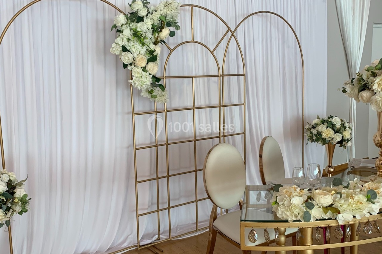 Décoration de mariage avec arche dorée, rideaux blancs, chaises élégantes et table ornée de fleurs blanches.