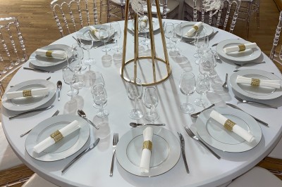 Table ronde élégamment dressée avec nappes blanches, assiettes, couverts, verres et serviettes ornées d'anneaux dorés.